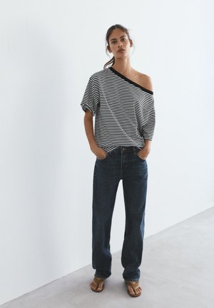 Žena nosiaca voľný čierno-biely pruhovaný top bez ramienok, tmavomodré džínsy a hnedé sandále stojí pri hladkej svetlej stene.