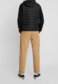 Czarna pikowana kamizelka z kapturem z poziomym przeszyciem, noszona na czarnej, długorękawowej koszulce, w połączeniu z beżowymi spodniami i czarnymi trampkami.