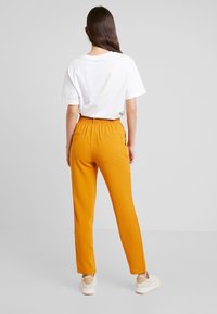 Oranga avsmalnande byxor i lätt tyg, med en elastisk midja, två sidofickor och en smidig yta, tillsammans med en vit T-shirt.