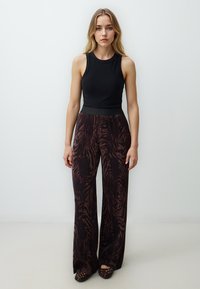 Schwarzes ärmelloses Oberteil mit hohem Kragen; weite, gemusterte dunkelbraune Hose mit strukturiertem, zebraähnlichem Design; flache schwarze gewebte Schuhe.