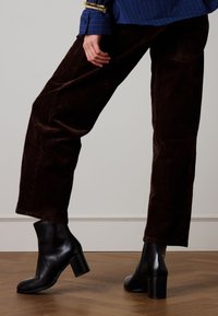 Black leather ankle boots with a chunky heel, paired with brown corduroy trousers. The outfit has a sleek, modern look with minimal accents.
