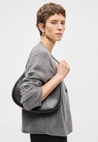 Bolsa de couro preta com forma curva e textura suave, segurada por uma pessoa com um cardigã cinza de lã, apresentando mangas largas e um botão.