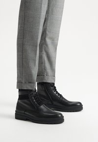 Czarne skórzane botki za kostkę z teksturowanym wierzchem, z zamknięciem na sznurowadło i zamek, połączone z szarymi spodniami w kratę, z mankietem na dole.