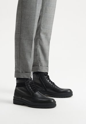 Czarne skórzane botki za kostkę z teksturowanym wierzchem, z zamknięciem na sznurowadło i zamek, połączone z szarymi spodniami w kratę, z mankietem na dole.