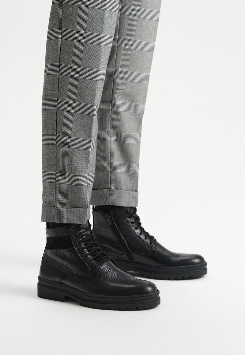 Czarne skórzane botki za kostkę z teksturowanym wierzchem, z zamknięciem na sznurowadło i zamek, połączone z szarymi spodniami w kratę, z mankietem na dole.