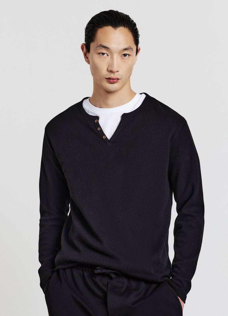 Pullover nero a maniche lunghe in tessuto testurizzato con scollatura a V, caratterizzato da tre bottoni marroni al colletto e una maglietta bianca sotto.