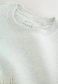 Svetlo šedý sveter s jemnou textúrou, rebrovaným výstrihom a potlačenou nálepkou s nápisom "PRE CELÚ RODINU." Obsahuje jemné farebné variácie.