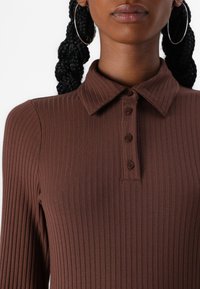 Brown ribbed polo shirt with a collar and four button placket. Features long sleeves and a textured fabric.