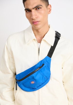 Blåt stoff crossbody-veske med en frontlomme med glidelås og logo; båret over en beige kordjakke mot en ensfarget bakgrunn.