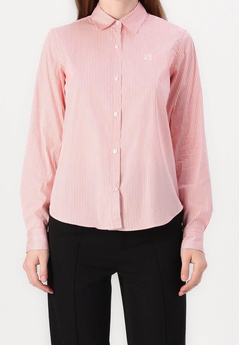 Femme portant une chemise à rayures verticales roses et blanches boutonnée, à manches longues, et un pantalon noir sur un fond blanc uni.