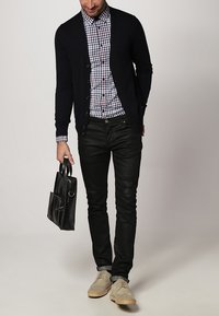 Marineblauwe cardigan over een geruit shirt, zwarte leren spijkerbroek, lichtbruine schoenen, en een zwarte leren tas met zijvak vasthoudend.