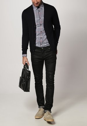 Granatowy kardigan na koszuli w kratę, czarne skórzane spodnie, jasnobeżowe buty, trzymający czarną skórzaną torbę z boczną kieszenią.