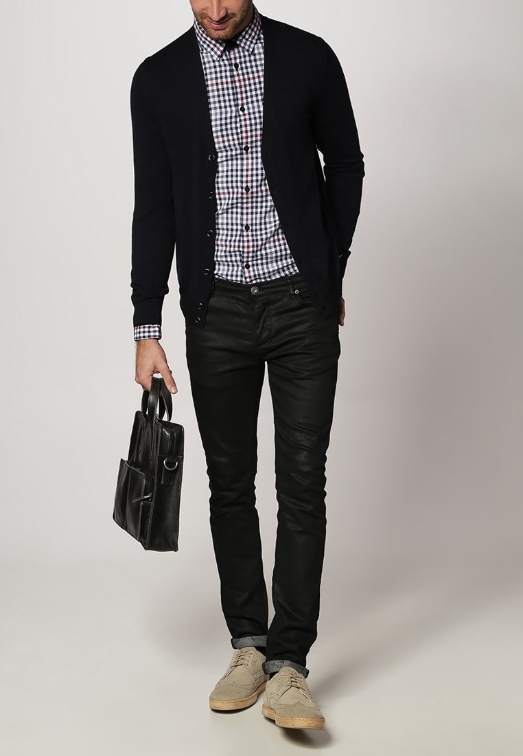 Marineblauwe cardigan over een geruit shirt, zwarte leren spijkerbroek, lichtbruine schoenen, en een zwarte leren tas met zijvak vasthoudend.