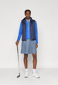 Conjunto de golfe com colete azul-marinho, camisa azul de mangas compridas e saia azul com padrão. Tênis brancos e um taco de golfe completam o visual.