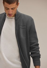 Cardigan en maille texturée gris foncé avec un col montant et une poche poitrine, porté sur un t-shirt gris clair. Comprend des poignets et un ourlet côtelés.