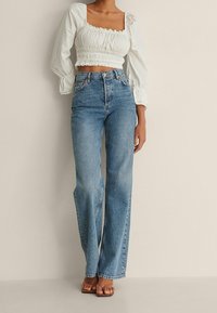 Kobieta w niebieskich dżinsach o wysokim stanie i prostych nogawkach, białej bluzce z marszczeniami na krótki rękaw oraz brązowych sandałach na obcasie z odkrytymi palcami.