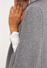 Grå stickad plagg med lös passform, med en texturerad yta och öppna sidor, i kombination med en vit spetsärm under.