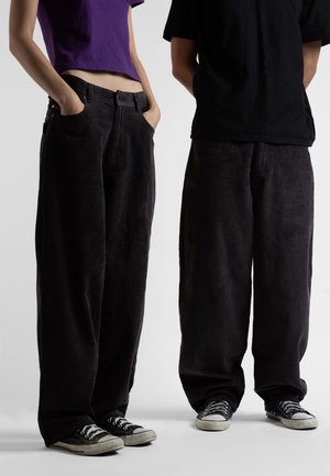 To personer iført løse sorte manchesterbukser og sorte sneakers, den ene med en lilla kort top og hænderne i lommerne.