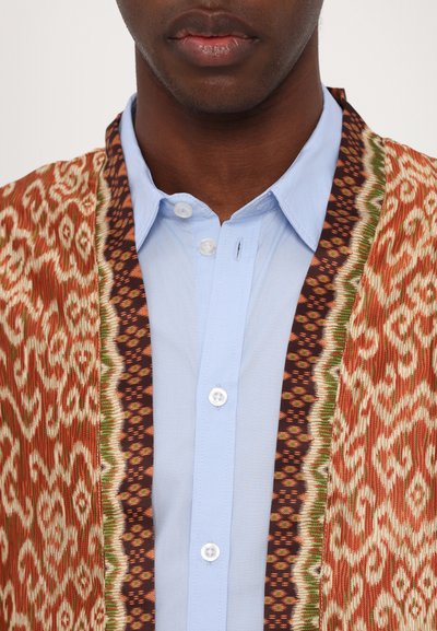 Chaqueta de tela multicolor con patrones intrincados, con un diseño naranja y blanco, superpuesta a una camisa de botón azul claro.