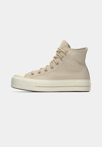 Beige høyhælte sneakers laget av glatt materiale. Har rund tå, seks snøringshull, gullfarget metallbeslag og en lavprofil, chunky såle.