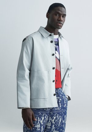 Jasnoszara kurtka z czarnymi guzikami, dwoma przednimi kieszeniami i kołnierzem. Pod spodem kolorowy sweter dzianinowy z abstrakcyjnymi wzorami.