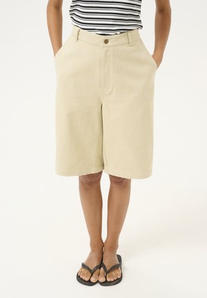 Knee-length beige shorts made of cotton fabric, featuring a front button, belt loops, and two side pockets. Worn with black flip-flops.