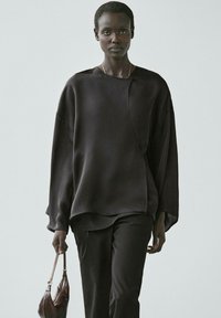 Top nero oversize in tessuto liscio, con scollo rotondo e orlo asimmetrico, abbinato a pantaloni scuri e una piccola borsa marrone.