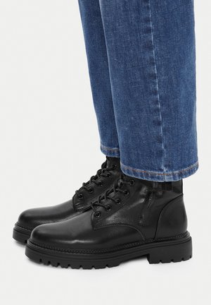 Schwarze Leder-Stiefeletten mit Schnürdesign, strukturierter Oberfläche und robuster Sohle, kombiniert mit blauen Jeans mit umgekrempeltem Saum.