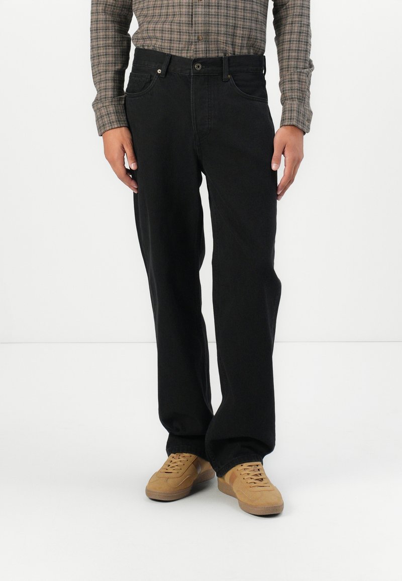 Jeans de mezclilla negros con un diseño de pierna recta, estilo de cinco bolsillos y cierre de botón. Llevados con una camisa de cuadros de manga larga y zapatillas color beige.