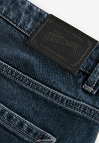 Tamno plave traperice s teksturiranom crnom kožnom etiketom na kojoj je uvučeno "Signature Denim Goods" i detaljnim šavovima duž šavova.