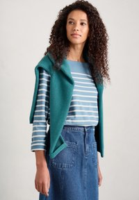 Tyrkysová pletená vesta přes modrou a bílou pruhovanou košili s dlouhým rukávem, doplněná sukní z denimu s vysokým pasem a dvěma zadními kapsami.