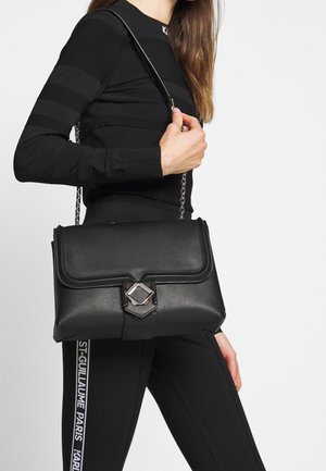 Sac à main en similicuir noir avec une forme structurée, doté d'un rabat, de ferrures argentées et d'une bandoulière chaîne, maintenu devant un fond blanc.