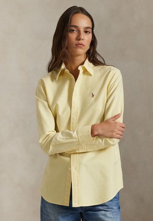 Kvinde med langt brunt hår iført en knappet lysegul langærmet skjorte og blå jeans, stående mod en ensfarvet beige baggrund.