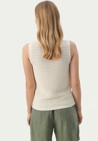 Kremowy bezrękawnik dzianinowy z teksturowanym wzorem, z prążkowanym dołem. Połączony z luźnymi zielonymi spodniami lnianymi, widok z tyłu.