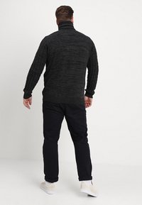 Pull en tricot noir à col roulé avec motif texturé ; poignets et ourlet côtelés ; porté avec un pantalon foncé et des chaussures claires, vu de dos.