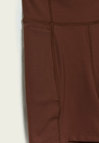 Ruskeat urheilushortsit, jotka on valmistettu sileästä kankaasta ja joissa on litteät saumat sekä sivutasku. Malli on yksinkertainen ja virtaviivainen.