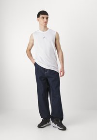 Hvid ærmeløs skjorte med sort logo, løse mørkeblå bukser og sorte sneakers. Enkel, afslappet stil med en løs pasform.