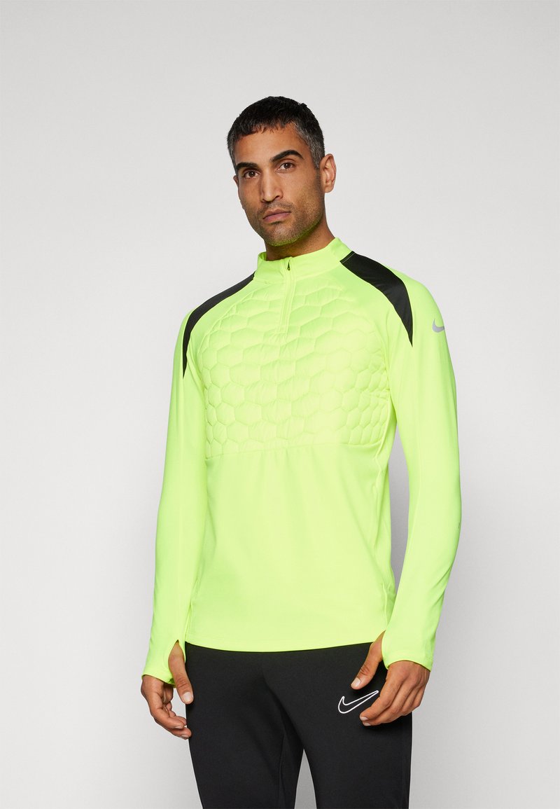 Mann, der ein neongelbes, langärmliges Sportoberteil mit hexagonalem Steppmuster und schwarzen Schulterpaneelen trägt, kombiniert mit schwarzen Nike-Hosen.