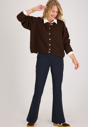 Vrouw die glimlacht en poseert, gekleed in een bruine gebuttonde trui over een witte shirt, marineblauwe flared broek en bruine sneakers met gumzolen.