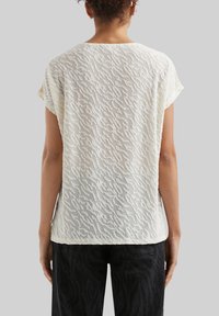 Top en crème léger avec un motif texturé en vagues, à manches courtes et à coupe décontractée. Le dos présente un design élégant et fluide.