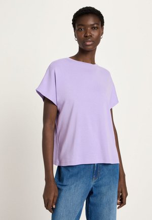 Woman with short hair wearing hoop earrings, a loose lilac short-sleeve top, and blue jeans, standing against a plain light background.