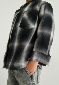 Sort og gråternet skjorte med en enkelt brystlomme, trykknapper og optrukne ærmer. Grå jeans med synlige syninger.