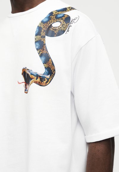 Λευκό πουκάμισο με κοντά μανίκια και μεγάλο πολύχρωμο γραφικό φιδιού τυλιγμένο κοντά στον ώμο, με ανοιχτό το στόμα και έξω τη γλώσσα, σε σκουρόχρωμο χέρι.