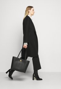 Black leather tote bag with gold hardware, smooth texture, and two long handles. Model wears a long black coat and high-heeled boots.