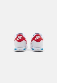 White Nike sneakers with red accents and a light blue heel, featuring a textured leather and mesh upper, visible branding on the heel.