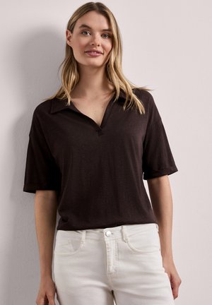 Femme aux cheveux blonds portant une chemise ample à manches courtes marron foncé avec col et un pantalon blanc taille haute, debout contre un mur clair.
