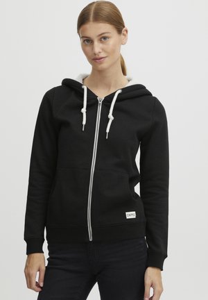 Juodas užtrauktukas hoody, pagamintas iš minkštos medvilnės, su balta dirbtinio kailio pamušalu, su raišteliais ir maža logotipo etikete apačioje.