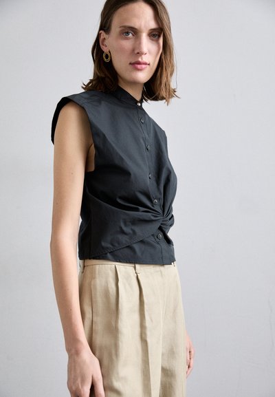 Cămașă fără mâneci, neagră, cu un design frontal răsucit, închidere cu buton și guler clasic; asociată cu pantaloni plisati bej deschis.