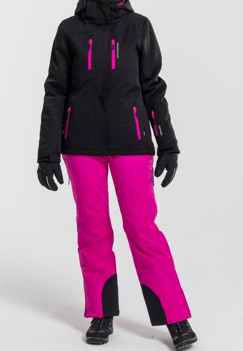 Femme portant une veste d'hiver noire avec des fermetures éclair roses, un pantalon de neige rose vif avec des patchs noirs, des gants noirs et des bottes noires.