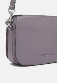 Helllila Leder-Crossbody-Tasche mit silbernen Beschlägen, Reißverschluss und geprägtem Logo-Detail. Glatte Textur und rechteckige Form.